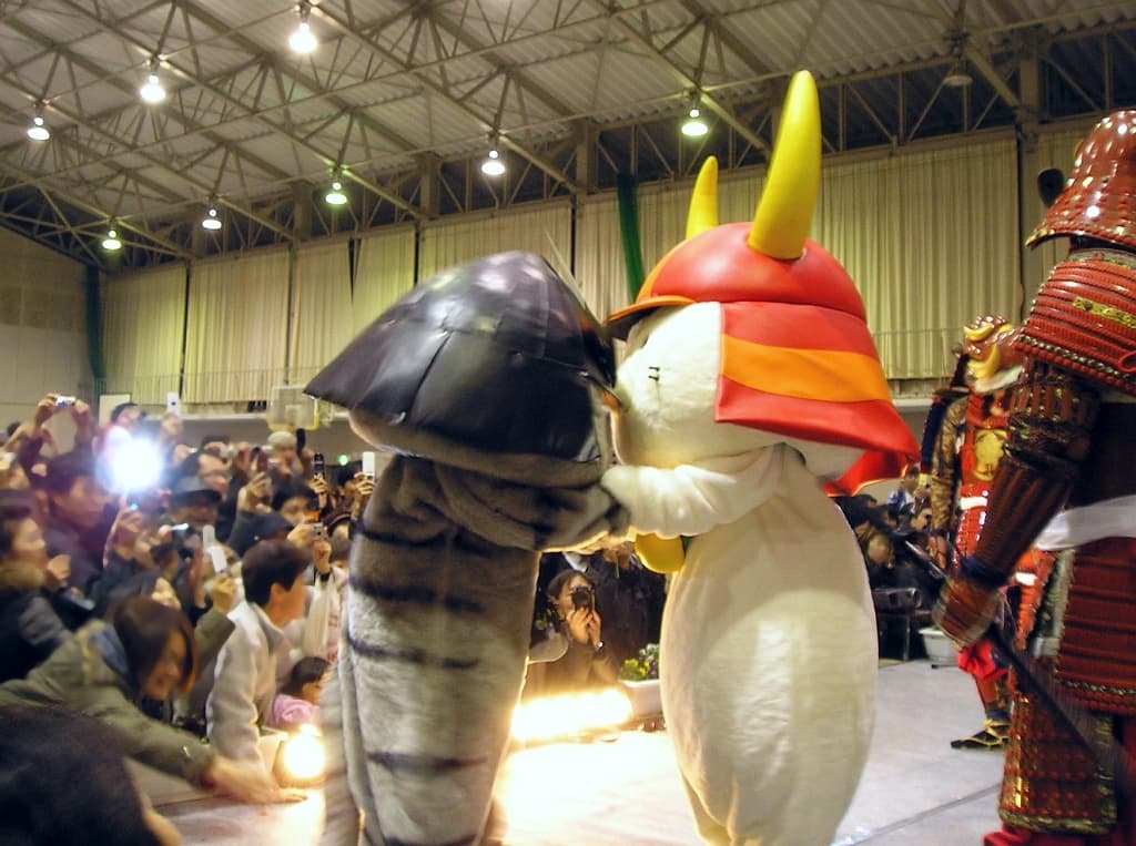 Hikonyan, a white cat mascot wearing a red samurai helmet with gold horns, greeting a tiger-costumed mascot at a public event