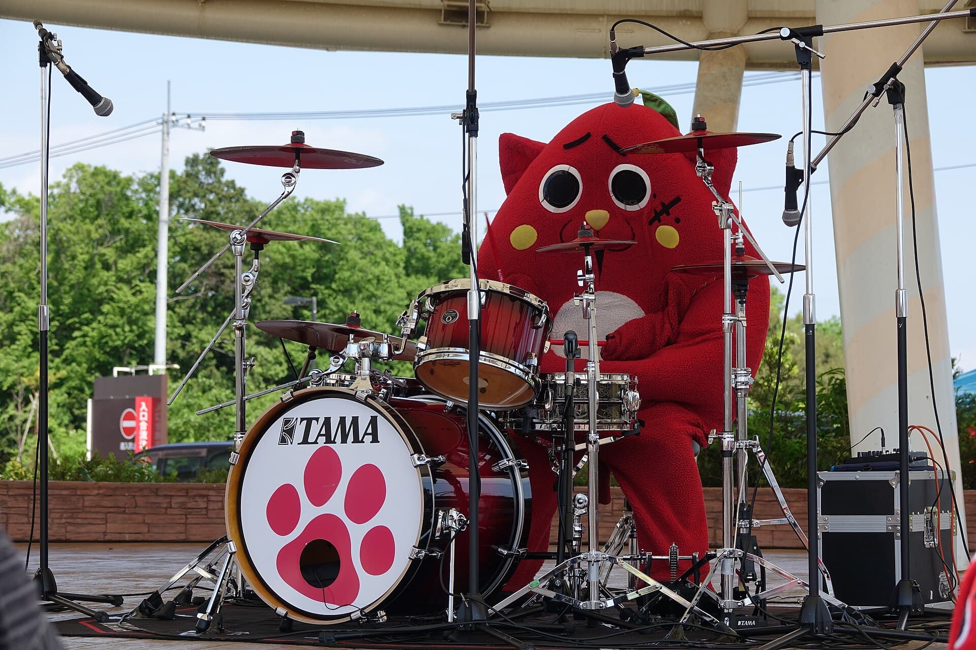 Nyango Star, a red cat-shaped mascot, playing a full drum kit on an outdoor stage